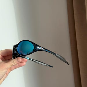 May include: Black and blue sunglasses with a marbled pattern on the frames and blue lenses. The temples are silver and gray. The sunglasses are held up in the air.