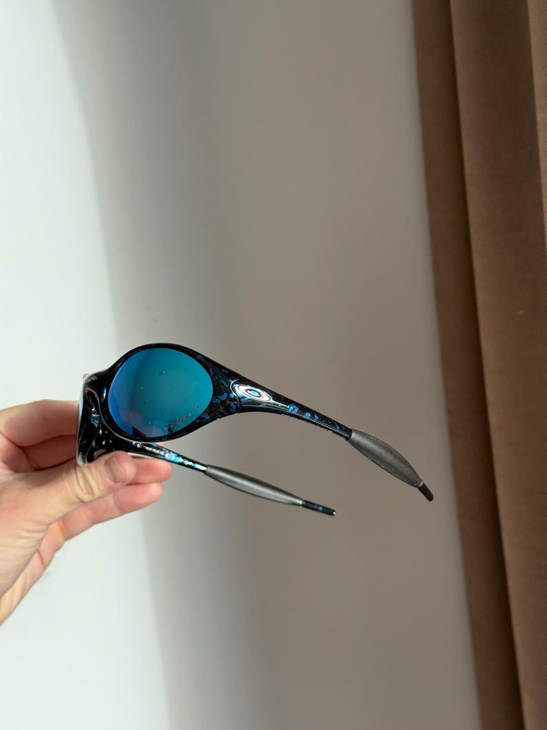 May include: Black and blue sunglasses with a marbled pattern on the frames and blue lenses. The temples are silver and grey. The sunglasses are held up in the air.
