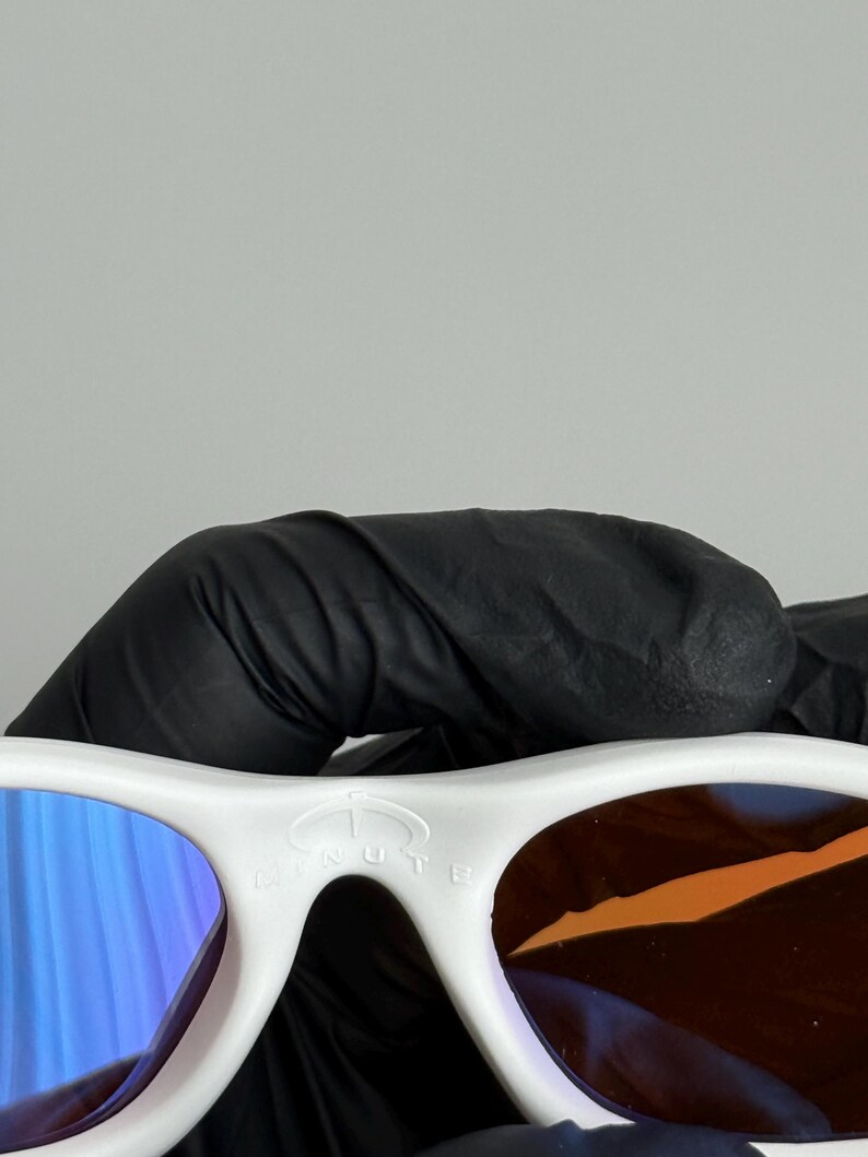 May include: White sunglasses with blue and brown lenses. The sunglasses have a white frame and the word "MINUTE" is printed on the top of the frame.