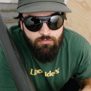 K&ouml;nnte beinhalten: Eine Person tr&auml;gt ein gr&uuml;nes T-Shirt mit dem Text "Libe Ide's", eine graue Kappe mit "metalwood" in Wei&szlig; und eine schwarze Sonnenbrille. Die Sonnenbrille hat einen dicken schwarzen Rahmen und verspiegelte Gl&auml;ser. Die Person hat einen Vollbart.