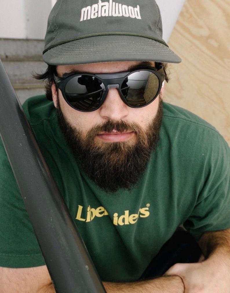 K&ouml;nnte beinhalten: Eine Person tr&auml;gt ein gr&uuml;nes T-Shirt mit dem Text "Libe Ide's", eine graue Kappe mit "metalwood" in Wei&szlig; und eine schwarze Sonnenbrille. Die Sonnenbrille hat einen dicken schwarzen Rahmen und verspiegelte Gl&auml;ser. Die Person hat einen Vollbart.