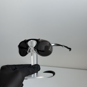 May include: Dark gray sunglasses featuring a distinct circular frame design with a bridge that connects at the top of the lenses. The temples are thin and metallic, and the lenses are dark, providing a sleek and functional appearance for eye protection.