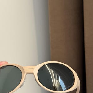 May include: Cream-colored sunglasses with dark lenses. The sunglasses have a rounded oval shape and a thick frame. The brand name "PIET" is printed on the top of the frame. The arms are thin and white.