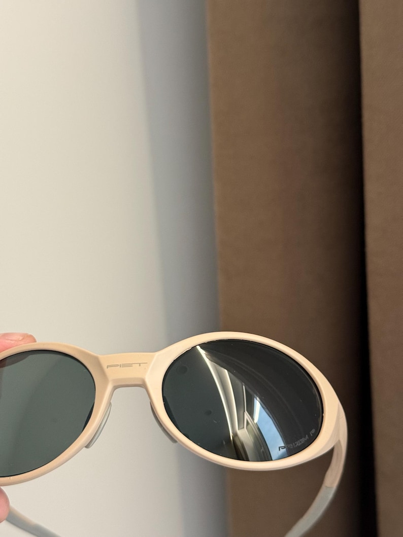 May include: Cream-colored sunglasses with dark lenses. The sunglasses have a rounded oval shape and a thick frame. The brand name "PIET" is printed on the top of the frame. The arms are thin and white.