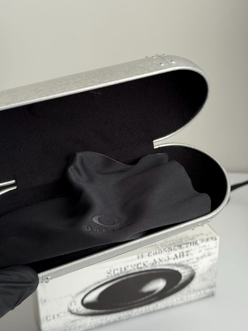 May include: Open silver-colored glasses case with a black interior and a black microfiber cloth. The case is oval-shaped with a riveted design. The cloth has the Oakley logo. A white box with text is partially visible.