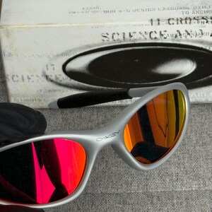 May include: Silver-framed Oakley sunglasses with red and orange mirrored lenses. The word "OAKLEY" is printed on the frame. The background includes a white box with the word "SCIENCE" and other text.