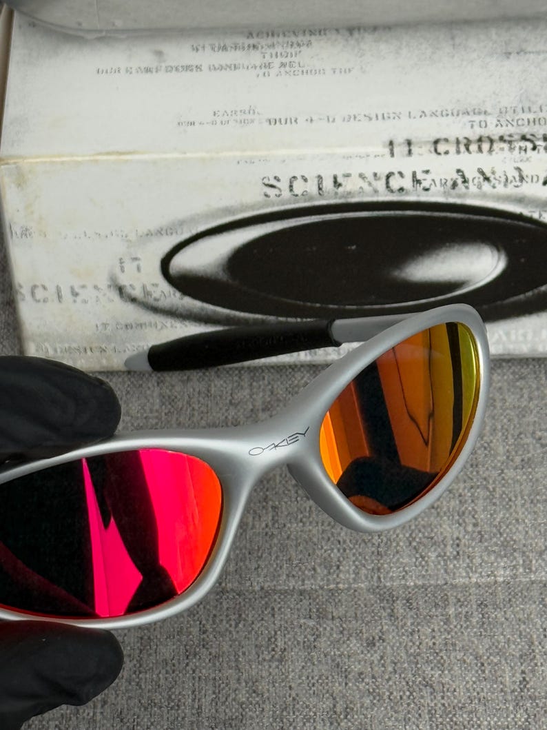 May include: Silver-framed Oakley sunglasses with red and orange mirrored lenses. The word "OAKLEY" is printed on the frame. The background includes a white box with the word "SCIENCE" and other text.