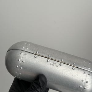 K&ouml;nnte beinhalten: Ein silberfarbenes, zylindrisches Etui mit einem Klappdeckel. Das Etui hat ein genietetes Design und der Text "CASE MADE IN CHINA" ist auf der Seite aufgedruckt. Das Etui wird von einer behandschuhten Hand gehalten.