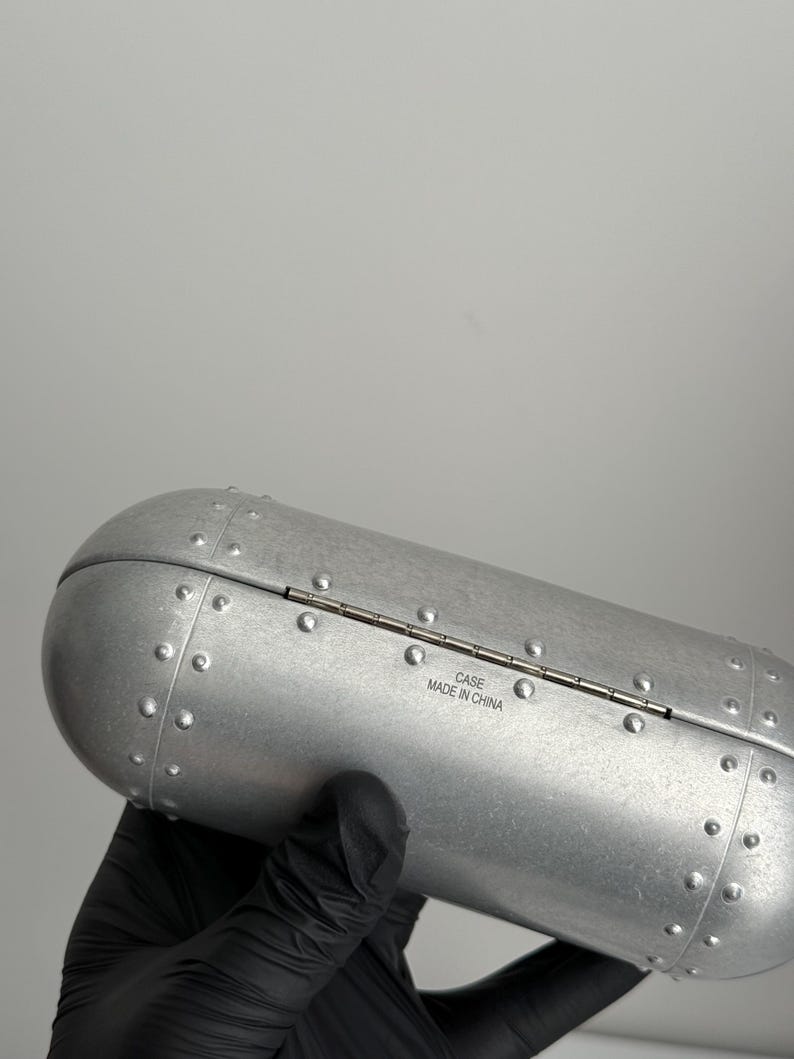 K&ouml;nnte beinhalten: Ein silberfarbenes, zylindrisches Etui mit einem Klappdeckel. Das Etui hat ein genietetes Design und der Text "CASE MADE IN CHINA" ist auf der Seite aufgedruckt. Das Etui wird von einer behandschuhten Hand gehalten.