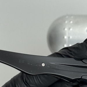 May include: A black object with text and a small silver accent. The object is held by a gloved hand. The background is a light gray color.