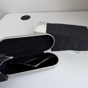May include: Open silver sunglasses case with black interior, holding dark sunglasses. A black microfiber cloth and a white box are next to the case. The sunglasses have a sleek, modern design.