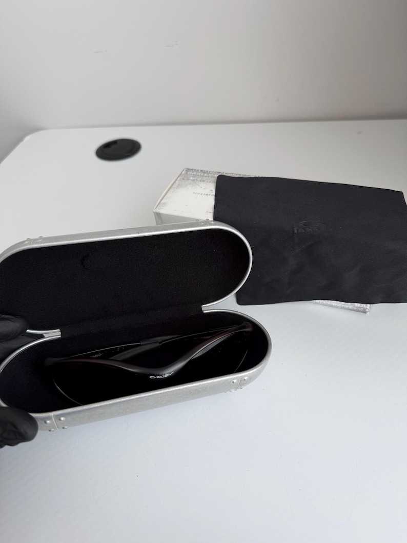 May include: Open silver sunglasses case with black interior, holding dark sunglasses. A black microfiber cloth and a white box are next to the case. The sunglasses have a sleek, modern design.