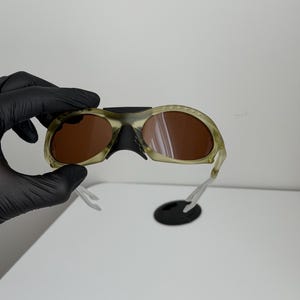 May include: Camouflage-patterned sunglasses with brown lenses. The frames are a mix of green and tan, with a black nose bridge and clear arms. A black, oval-shaped piece is attached to the sunglasses.