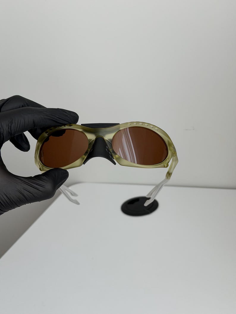 May include: Camouflage-patterned sunglasses with brown lenses. The frames are a mix of green and tan, with a black nose bridge and clear arms. A black, oval-shaped piece is attached to the sunglasses.