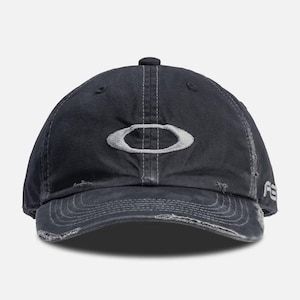 Op de afbeelding: Een zwarte baseballcap met een distressed afwerking, met het witte Oakley-logo. De cap heeft een gebogen klep en een versleten uitstraling. De stiksels zijn zichtbaar en de stijl suggereert een vintage of voorgewassen esthetiek.