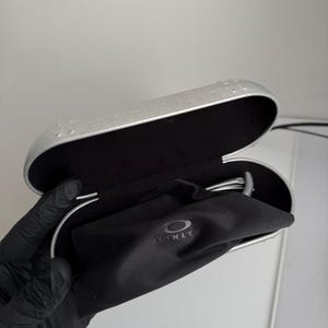 Puede incluir: Estuche de gafas plateado abierto con interior negro y una bolsa negra con cord&oacute;n. La bolsa tiene el nombre de la marca "OAKLEY" impreso. El estuche est&aacute; abierto, revelando el contenido.