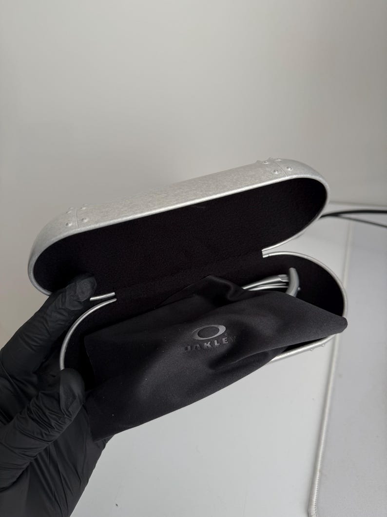 Puede incluir: Estuche de gafas plateado abierto con interior negro y una bolsa negra con cord&oacute;n. La bolsa tiene el nombre de la marca "OAKLEY" impreso. El estuche est&aacute; abierto, revelando el contenido.