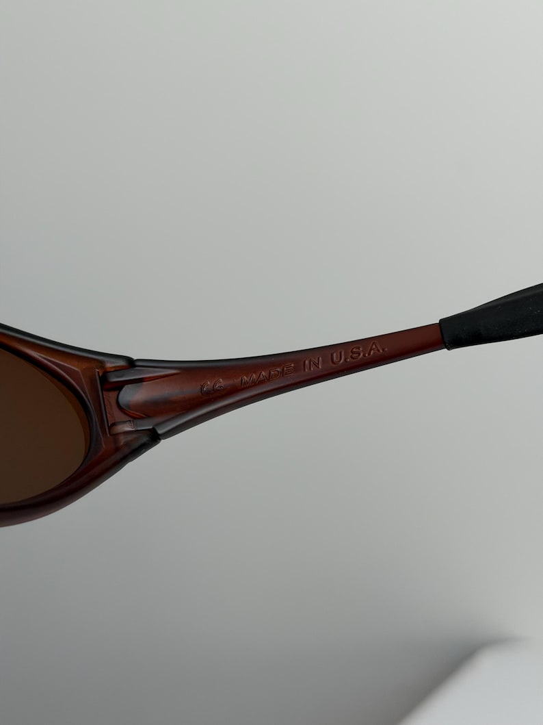 May include: Close-up of brown sunglasses with dark lenses. The frame is a dark brown color with the text "MADE IN U.S.A." printed on the arm.