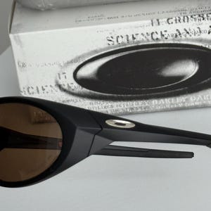 Puede incluir: Gafas de sol negras con lentes marrones. Las gafas de sol tienen un dise&ntilde;o elegante y moderno con un acabado mate. La montura presenta un peque&ntilde;o acento plateado cerca de la patilla. Las gafas de sol est&aacute;n delante de una caja blanca con el texto "SCIENCE AND".