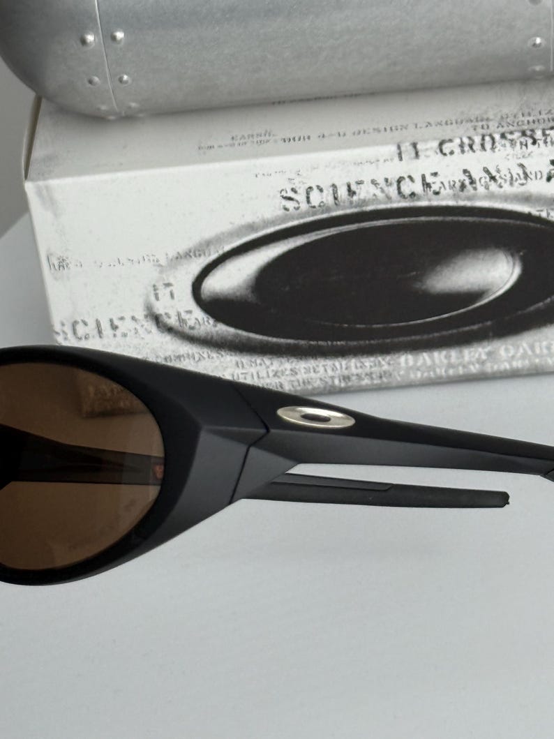 Puede incluir: Gafas de sol negras con lentes marrones. Las gafas de sol tienen un dise&ntilde;o elegante y moderno con un acabado mate. La montura presenta un peque&ntilde;o acento plateado cerca de la patilla. Las gafas de sol est&aacute;n delante de una caja blanca con el texto "SCIENCE AND".