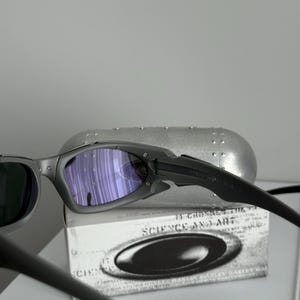 May include: Gray sunglasses with purple-tinted lenses and black arms. The sunglasses are resting on a white box with the words "SCIENCE AND ART" printed on it. A silver case is in the background.