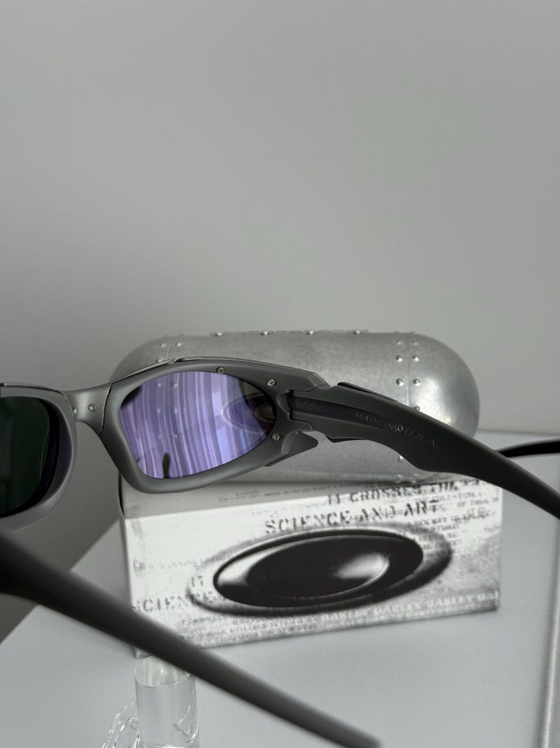 May include: Gray sunglasses with purple-tinted lenses and black arms. The sunglasses are resting on a white box with the words "SCIENCE AND ART" printed on it. A silver case is in the background.