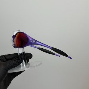 May include: Purple sunglasses with red lenses. The frames are a vibrant purple with black accents on the temples. The sunglasses are displayed on a clear stand against a plain background.