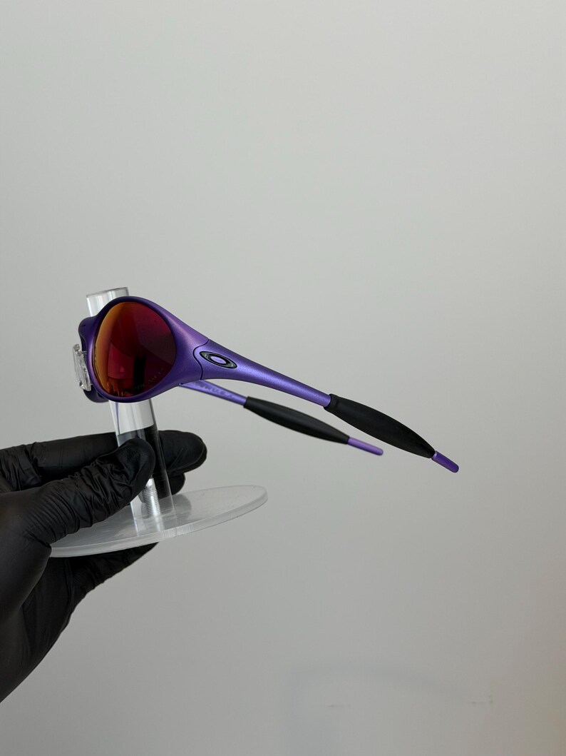 May include: Purple sunglasses with red lenses. The frames are a vibrant purple with black accents on the temples. The sunglasses are displayed on a clear stand against a plain background.