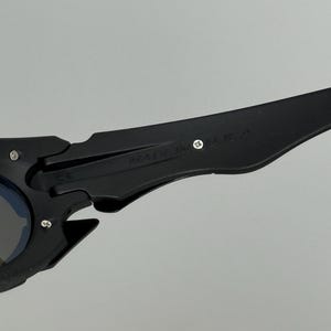 May include: Black sunglasses with a unique, angular design. The frame features a matte finish and the text "MADE IN U.S.A." is visible on the arm. The lenses have a blue tint.