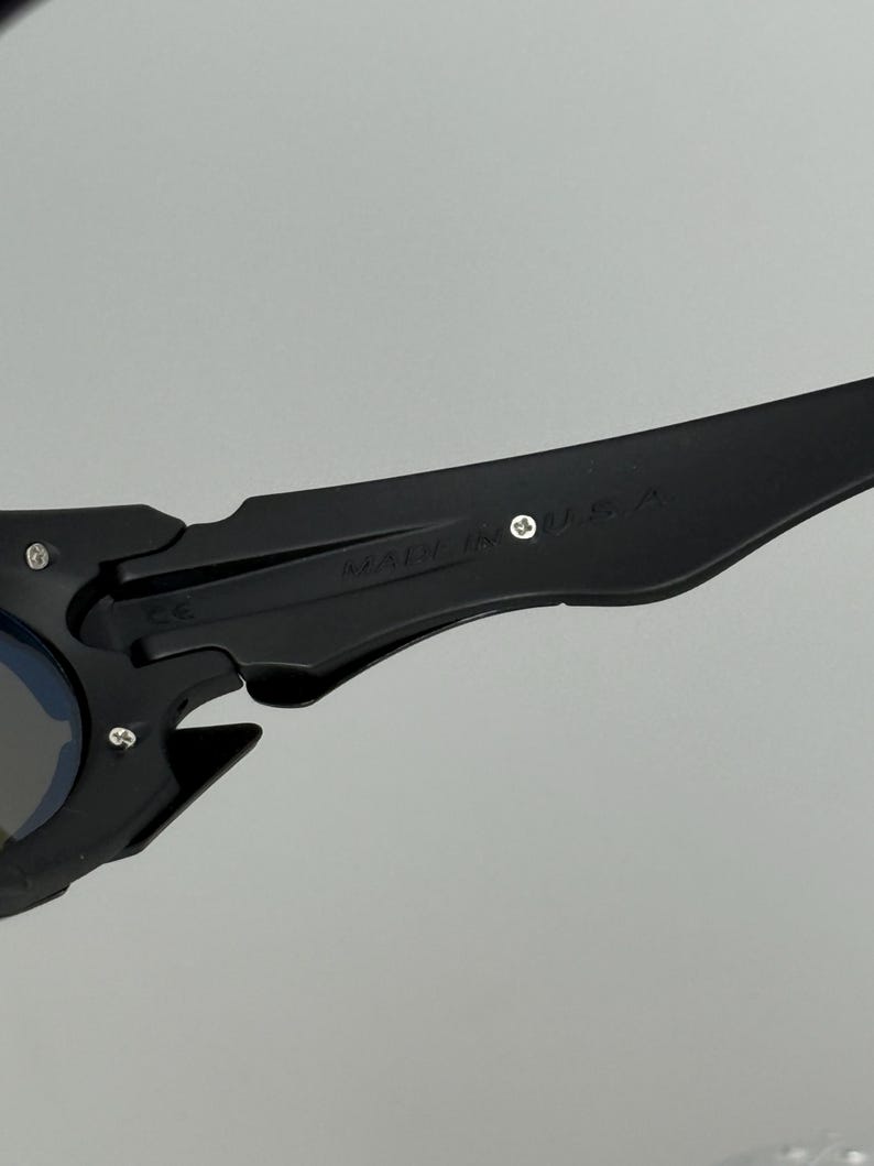 May include: Black sunglasses with a unique, angular design. The frame features a matte finish and the text "MADE IN U.S.A." is visible on the arm. The lenses have a blue tint.