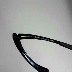 May include: Close-up of eyeglasses with a black frame and blue patterned accents. The text "OO9450-1259 59 22 133 3N" is printed on the frame. The glasses have a sleek, modern design.