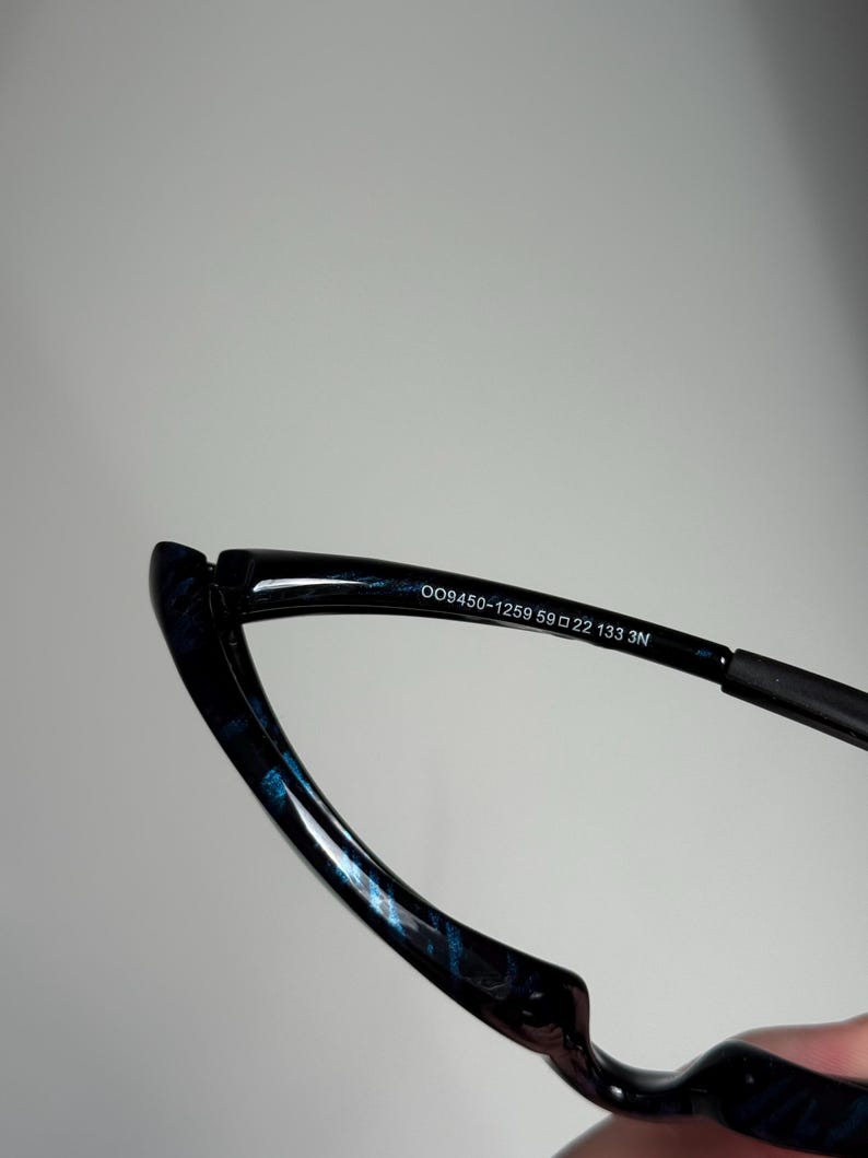 May include: Close-up of eyeglasses with a black frame and blue patterned accents. The text "OO9450-1259 59 22 133 3N" is printed on the frame. The glasses have a sleek, modern design.