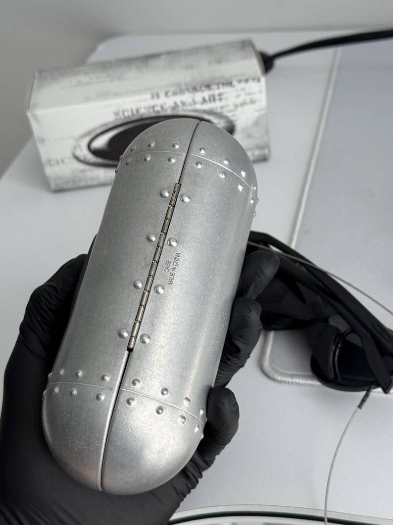 K&ouml;nnte beinhalten: Ein silberfarbenes, genietetes Brillenetui mit Scharnier&ouml;ffnung. Das zylindrische Etui hat im Hintergrund eine Schachtel mit dem Text "SCIENCE AND ART". Das Etui wird von einer Person mit schwarzen Handschuhen gehalten. Hergestellt in China.