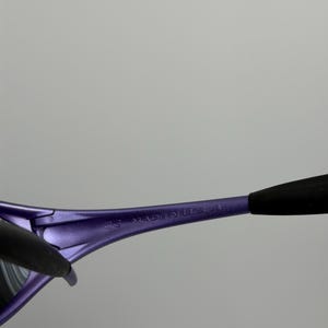 May include: Close-up of purple sunglasses with black arms. The text "MADE IN U.S.A." is printed on the purple frame. The sunglasses have a sleek, modern design.