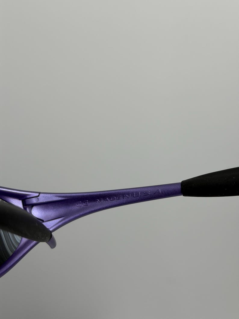 May include: Close-up of purple sunglasses with black arms. The text "MADE IN U.S.A." is printed on the purple frame. The sunglasses have a sleek, modern design.