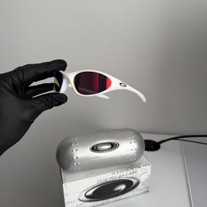 May include: White sunglasses with red lenses and a silver case. The sunglasses feature a streamlined design with curved arms and a bold frame. The case is metallic and has a rounded shape with the brand's logo. The box below the case has the text "SCIENCE AND ART".