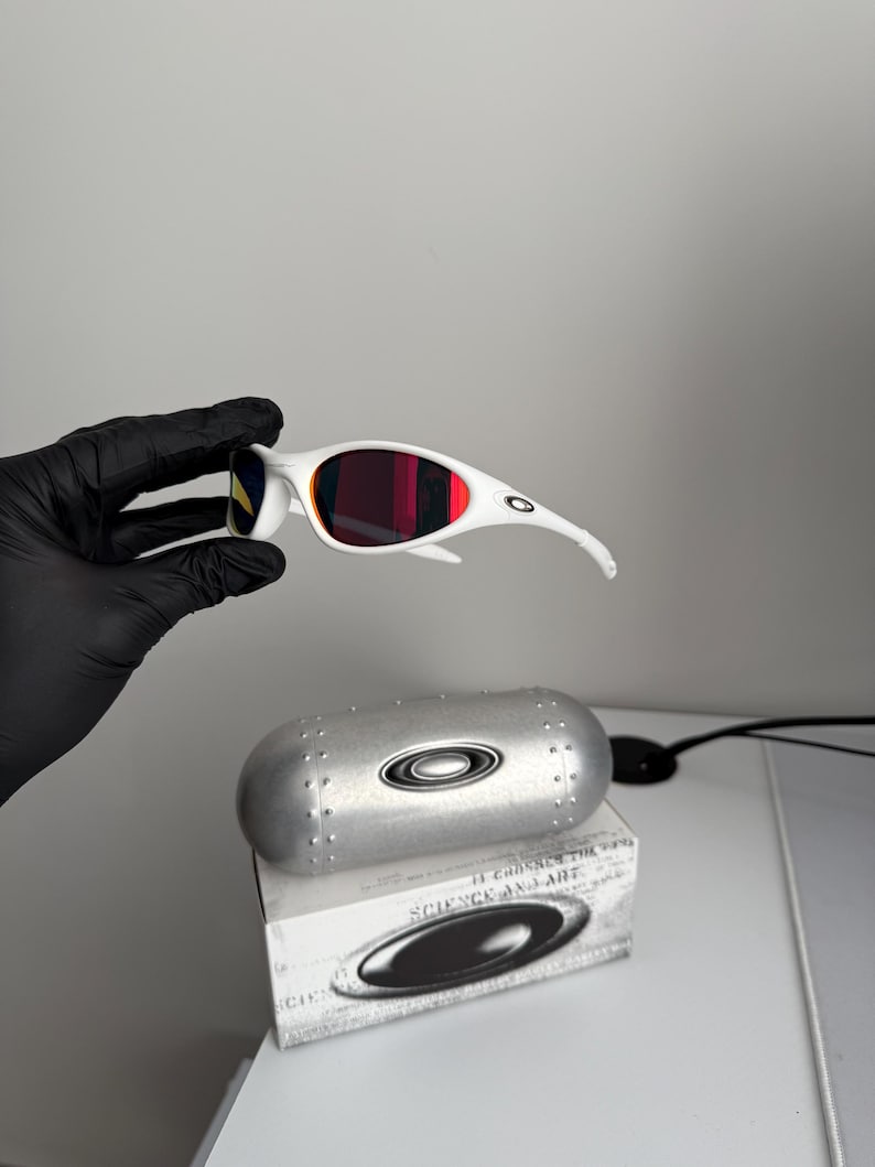 May include: White sunglasses with red lenses and a silver case. The sunglasses feature a streamlined design with curved arms and a bold frame. The case is metallic and has a rounded shape with the brand's logo. The box below the case has the text "SCIENCE AND ART".