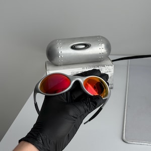 Pu&ograve; includere: Occhiali da sole con montatura argentata e lenti rosse. Gli occhiali sono tenuti da una mano guantata, con un astuccio argentato e una scatola sullo sfondo. Le lenti hanno un effetto sfumato, dal rosso all'arancione.