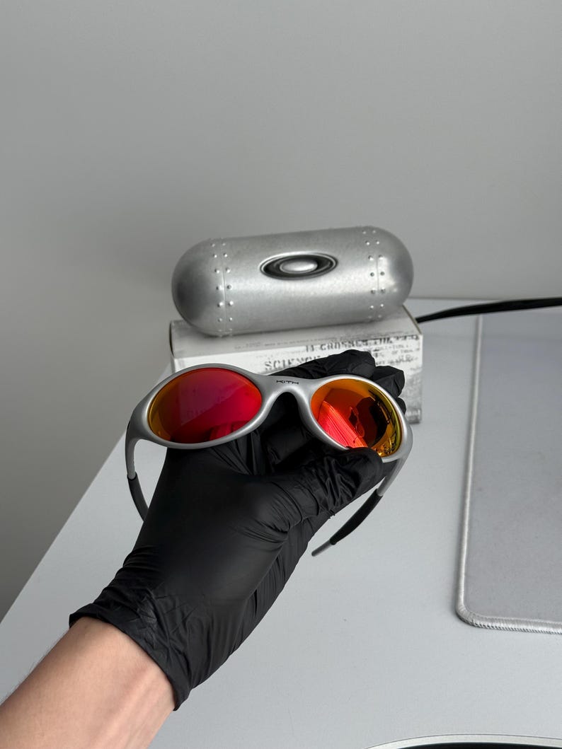 Pu&ograve; includere: Occhiali da sole con montatura argentata e lenti rosse. Gli occhiali sono tenuti da una mano guantata, con un astuccio argentato e una scatola sullo sfondo. Le lenti hanno un effetto sfumato, dal rosso all'arancione.
