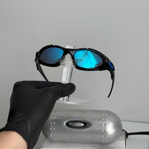 May include: Black framed sunglasses with blue mirrored lenses are displayed on a clear stand. The sunglasses are set against a white background, with a silver metal case and a box with the text "SCIENCE AND ART" visible.