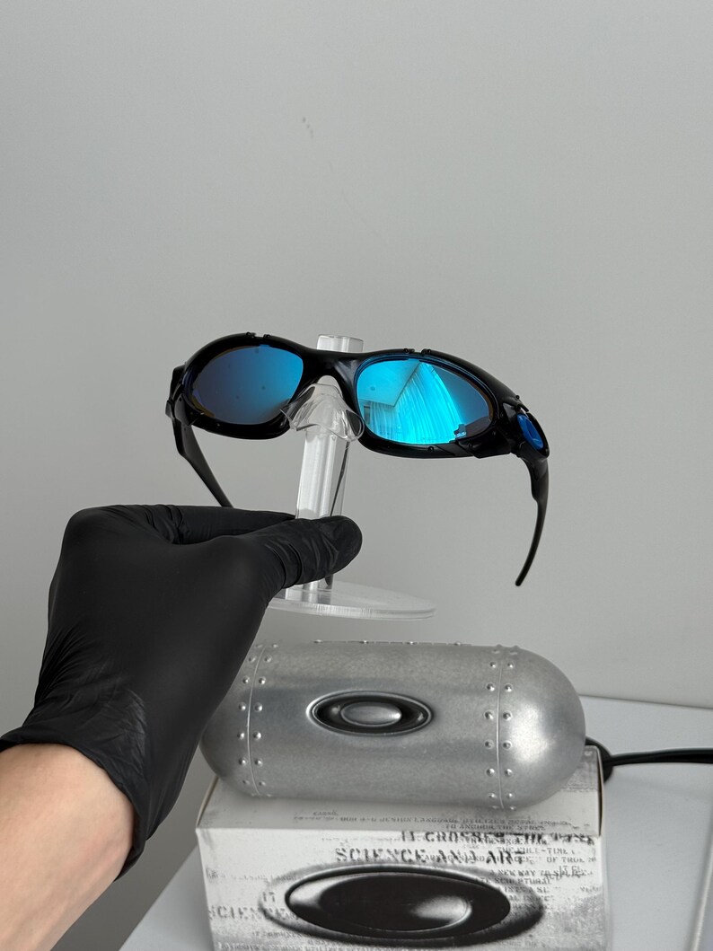May include: Black framed sunglasses with blue mirrored lenses are displayed on a clear stand. The sunglasses are set against a white background, with a silver metal case and a box with the text "SCIENCE AND ART" visible.