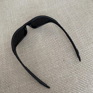 Puede incluir: Gafas de sol negras con un dise&ntilde;o elegante y moderno. Las gafas tienen una montura rectangular y lentes oscuras. Las patillas son finas y se curvan ligeramente, terminando en una punta. Las gafas descansan sobre una superficie clara y texturizada.