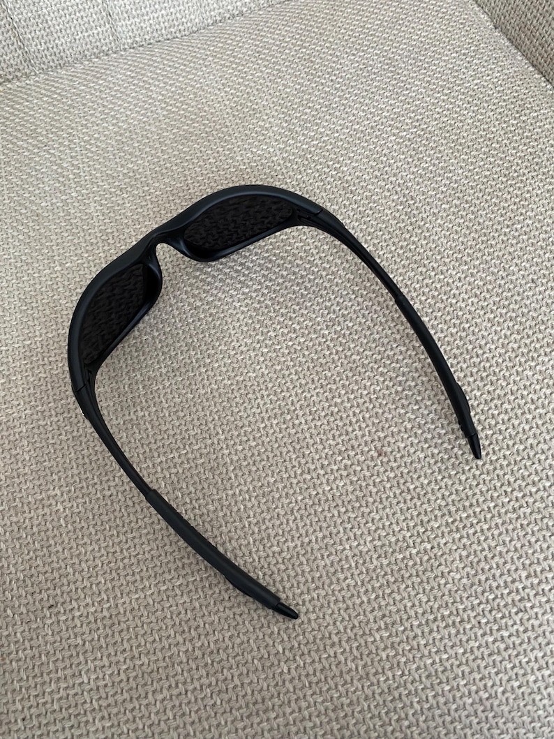 Puede incluir: Gafas de sol negras con un dise&ntilde;o elegante y moderno. Las gafas tienen una montura rectangular y lentes oscuras. Las patillas son finas y se curvan ligeramente, terminando en una punta. Las gafas descansan sobre una superficie clara y texturizada.