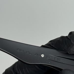 May include: Close-up of a black plastic object with text and a screw. The object is held by a gloved hand. The background is a plain, light gray.