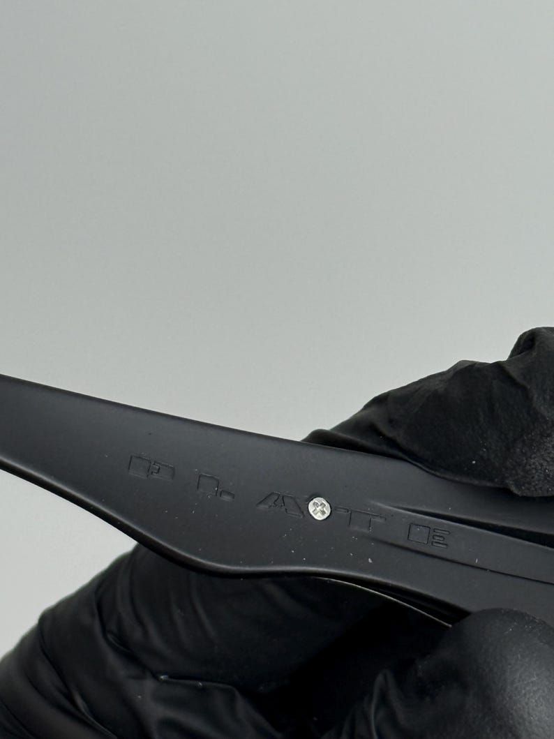 May include: Close-up of a black plastic object with text and a screw. The object is held by a gloved hand. The background is a plain, light gray.
