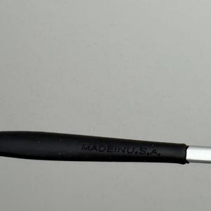 Puede incluir: Primer plano de un mango negro con el texto "MADE IN U.S.A." en blanco. El mango parece estar hecho de goma o un material similar, y est&aacute; unido a un objeto met&aacute;lico plateado.
