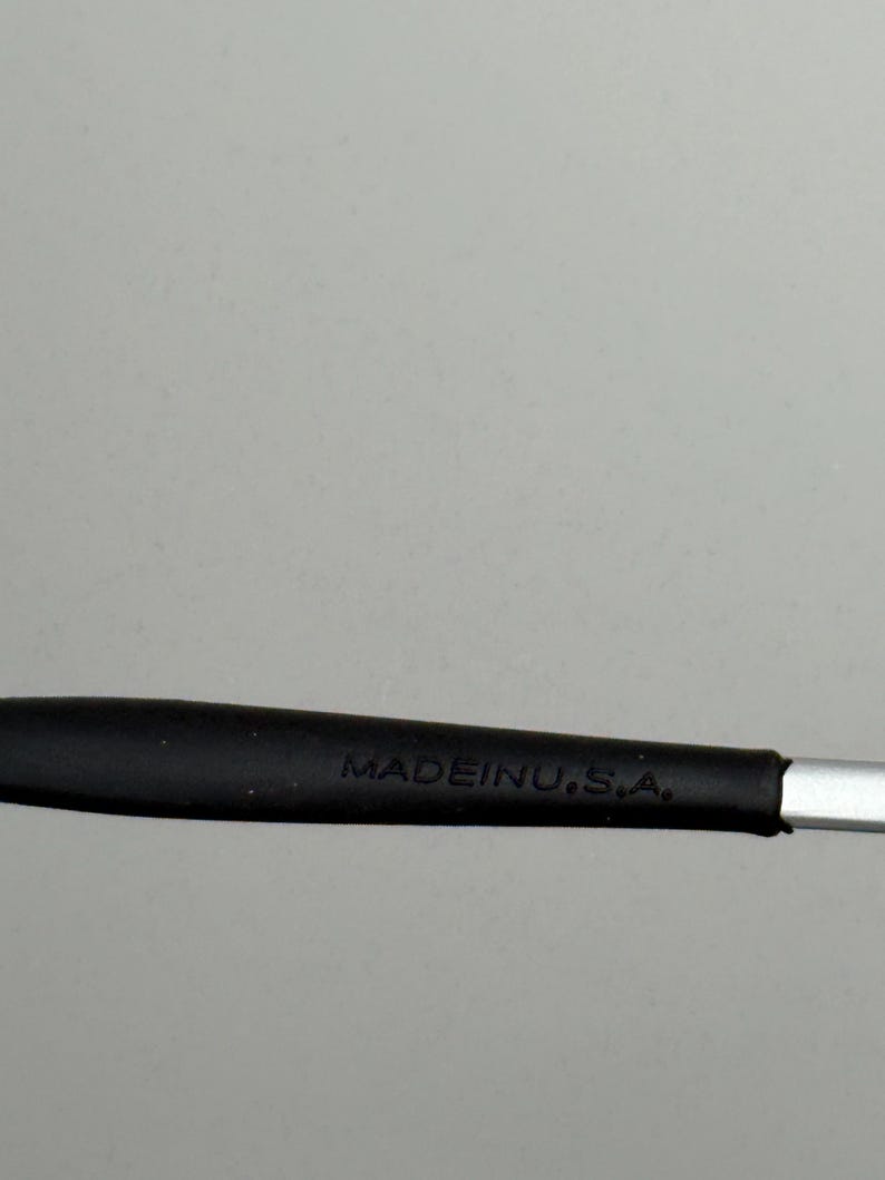 Puede incluir: Primer plano de un mango negro con el texto "MADE IN U.S.A." en blanco. El mango parece estar hecho de goma o un material similar, y est&aacute; unido a un objeto met&aacute;lico plateado.