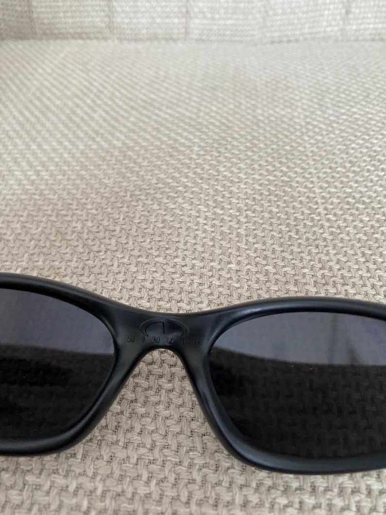 Puede incluir: Gafas de sol negras con lentes oscuras. Las gafas de sol tienen un dise&ntilde;o elegante y curvado y un acabado mate. La montura presenta un logotipo sutil en el puente. Las lentes parecen estar tintadas, ofreciendo protecci&oacute;n solar.