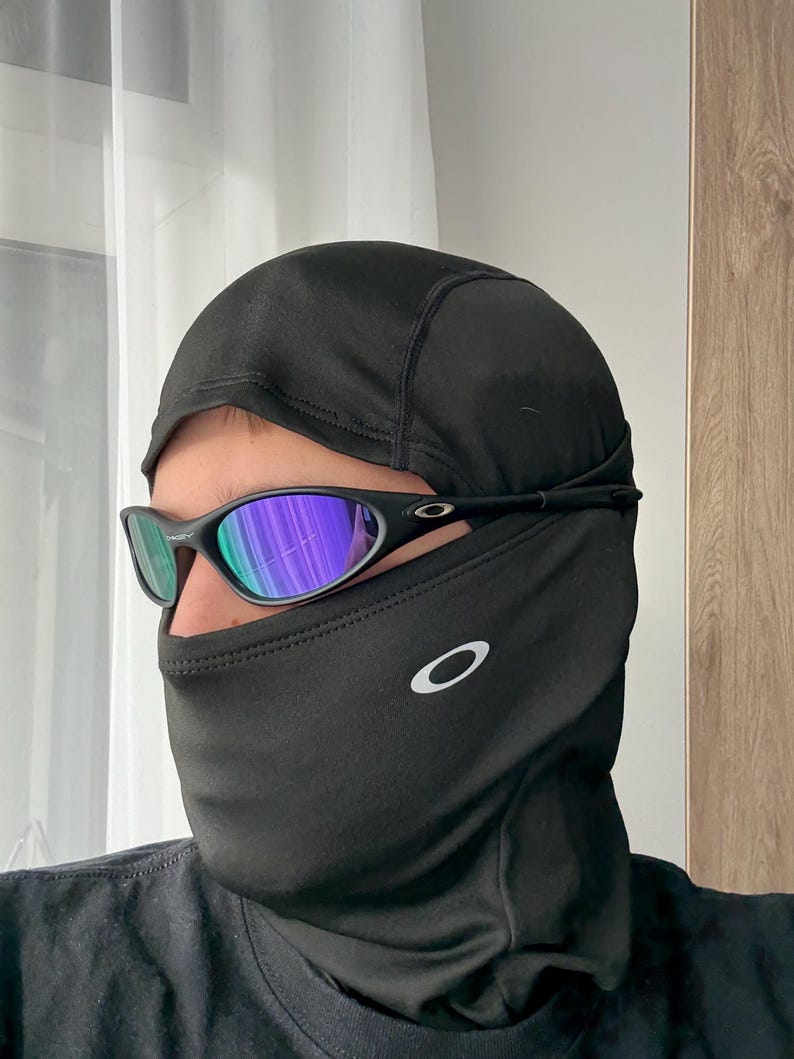 May include: Black balaclava and sunglasses. The balaclava covers the head and neck, with a small white logo on the side. The sunglasses have a black frame and reflective lenses with blue and green hues.