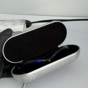 May include: Open silver sunglasses case with black interior and black sunglasses. The case has a riveted design and a silver trim. The sunglasses are black with a blue reflection.